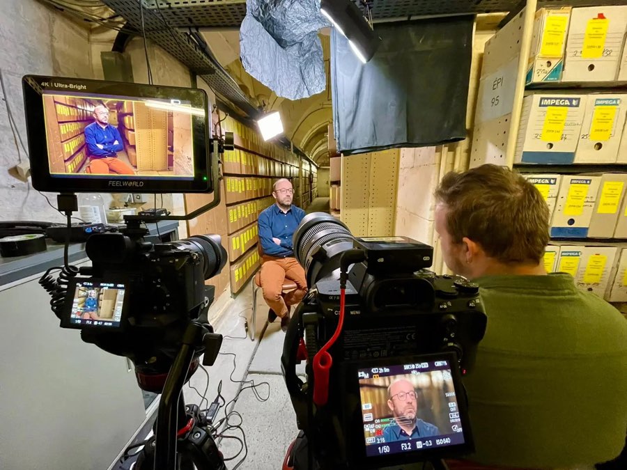 Tournage d'une interview dans un local d'archives : deux caméras cadrent un homme assis entre des rayonnages de boîtes.
